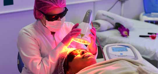 Profissional de estética em ambiente clínico, vestindo jaleco rosa, touca, máscara e óculos de proteção escuros, aplica equipamento de laser ou LED no rosto de uma paciente deitada em maca. A luz intensa alaranjada do dispositivo ilumina o rosto da cliente. Equipamentos eletrônicos ao fundo.