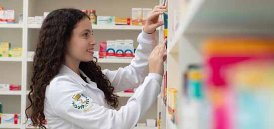 Estudante feminina de Farmácia, com jaleco branco com brasão institucional e cabelo encaracolado longo, sorri enquanto organiza ou busca medicamentos em prateleiras bem abastecidas de uma farmácia escola. Caixas coloridas de remédios de diferentes tamanhos preenchem as prateleiras ao fundo.