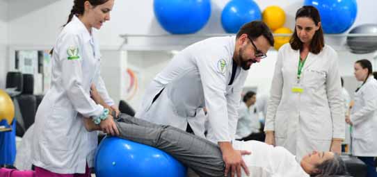 Sessão de fisioterapia em clínica escola universitária. Um fisioterapeuta masculino com jaleco branco e óculos realiza técnica de reabilitação com bola de exercício azul grande em uma paciente deitada no chão. Duas estudantes femininas de jaleco branco observam e auxiliam o procedimento. Bolas coloridas de pilates ao fundo.