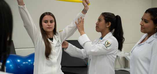 Três estudantes de Fisioterapia em jalecos brancos com brasão institucional realizam avaliação funcional de membro superior. Uma paciente/estudante com o braço levantado acima da cabeça é avaliada por colega que posiciona uma faixa elástica amarela para teste de amplitude de movimento. Uma terceira estudante observa o procedimento atentamente. Ao fundo, bola de pilates azul e equipamentos de reabilitação compõem a clínica escola.