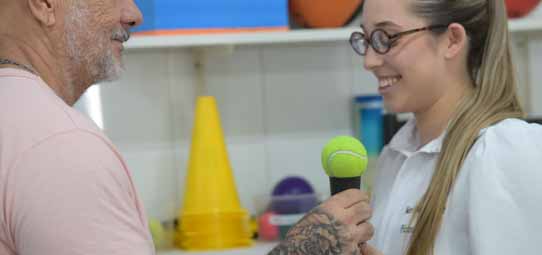 Estudante feminina de Fisioterapia, de jaleco branco, cabelo loiro comprido e óculos de armação grossa, sorri enquanto realiza atendimento com paciente idoso de barba branca e camiseta rosa. Ela segura uma bola de tênis adaptada como cabo de microfone em atividade lúdica ou de reabilitação fonoarticulatória. O paciente observa o equipamento improvisado com atenção. Ao fundo, cones amarelos, bolas coloridas e materiais de fisioterapia compõem a clínica escola.