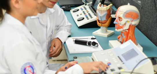 Estudante de Fonoaudiologia em jaleco branco opera equipamento de audiometria sobre bancada com modelos anatômicos da laringe, cabeça com músculos faciais e instrumentos de avaliação. O ambiente é um laboratório clínico com aparelhos especializados para diagnóstico auditivo e vocal.