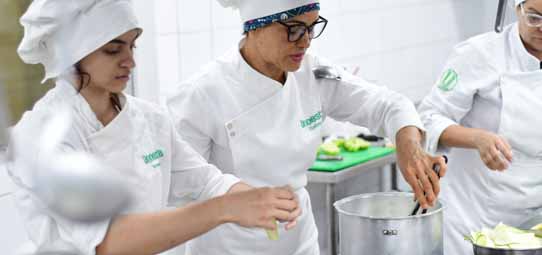 Três estudantes de Gastronomia com uniformes brancos — dólmã, avental e touca — trabalham em cozinha profissional. Um deles mexe o conteúdo de uma panela grande de aço inox enquanto outro manuseia ingredientes vegetais verdes. A bancada de aço inox e equipamentos de cozinha profissional compõem o cenário.