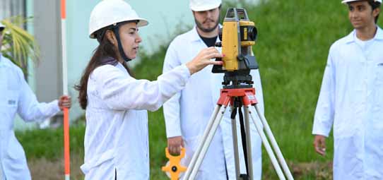 Estudante feminina de cabelo longo, capacete branco e jaleco branco opera uma estação total amarela da marca Leica sobre tripé em área externa gramada do campus. Ao fundo, outros dois estudantes com equipamentos observam a atividade prática de topografia e geoprocessamento.