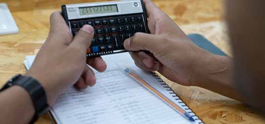 Mãos masculinas seguram uma calculadora financeira HP 12C sobre uma mesa com caderno de anotações e lista de números. O visor da calculadora exibe o número 2534225.84. Um relógio de pulso preto aparece no punho esquerdo. A cena representa cálculos financeiros e gestão comercial.