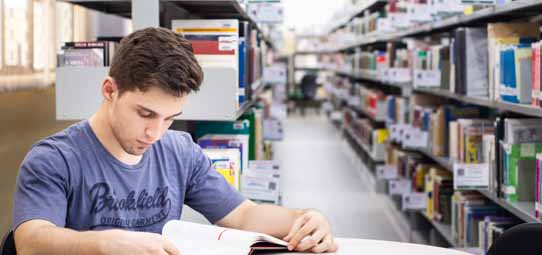 Jovem estudante masculino, de camiseta azul com estampa "Brookfield", sentado em mesa de estudos em uma biblioteca universitária, lendo um livro aberto com atenção. Ao fundo, fileiras de estantes com livros de diversas cores se estendem criando perspectiva de profundidade.