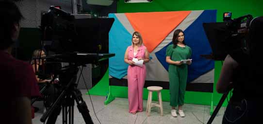 Estúdio de televisão universitário com duas apresentadoras posicionadas diante de um cenário colorido com formas geométricas em laranja, verde e azul. Uma está de pé usando calça e blusa rosa e a outra de blusa verde, ambas segurando roteiros. Câmeras profissionais em primeiro plano e teleprompter visível.