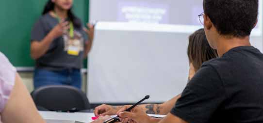 Sala de aula universitária com alunos sentados em carteiras. Uma estudante de pé ao fundo, de camiseta cinza e crachá, apresenta conteúdo projetado em tela branca. Em primeiro plano, um estudante masculino de camiseta preta toma notas com caneta. A imagem representa atividade de apresentação oral em aula de Letras.
