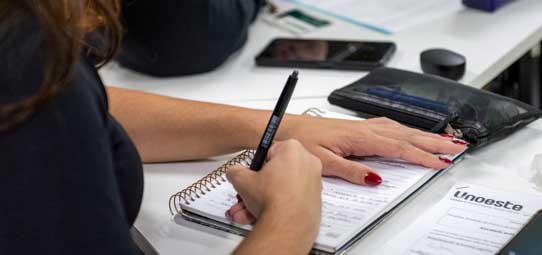 Aluna escreve com caneta em material impresso apoiado sobre um caderno durante atividade acadêmica em sala de aula. Sobre a mesa aparecem estojo, celular e outros materiais de estudo, enquanto a estudante realiza anotações no conteúdo.