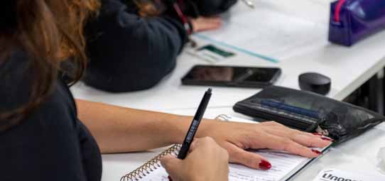 Close na mão direita de uma estudante com unhas vermelhas, escrevendo em caderno espiral com caneta preta sobre mesa branca de sala de aula. Ao fundo desfocado, uma pasta preta, smartphone e material de outro aluno.