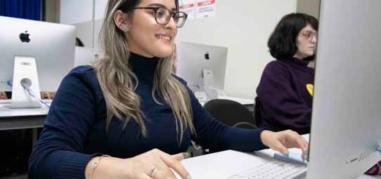 Estudante feminina sorridente, de óculos redondos, cabelo loiro e suéter azul marinho, trabalha em um computador iMac branco em laboratório de informática. Ao fundo, outra estudante de suéter roxo/bordô trabalha em seu iMac. O ambiente remete ao laboratório de Marketing com computadores Apple.
