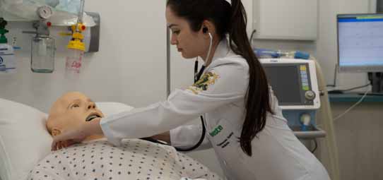 Estudante feminina de Medicina, com jaleco branco com logotipo Unoeste e cabelo escuro preso, usa estetoscópio para auscultar manequim de simulação clínica de adulto deitado em maca hospitalar. A estudante se inclina levemente sobre o manequim com postura técnica e expressão concentrada. Ao fundo, aparelhos hospitalares, monitor e equipamento de ventilação compõem o laboratório de simulação realística.