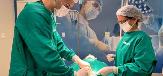 Dois estudantes de Medicina com scrubs verdes, máscaras cirúrgicas, toucas e luvas realizam procedimento cirúrgico simulado em manequim coberto com campos cirúrgicos verdes. O estudante masculino à esquerda utiliza instrumento cirúrgico enquanto a estudante feminina de óculos à direita auxilia o procedimento. Ao fundo, um painel fotográfico realista representa uma sala cirúrgica completa com equipe operando, criando imersão no ambiente hospitalar. As roupas têm o logotipo "Unoeste" visível.