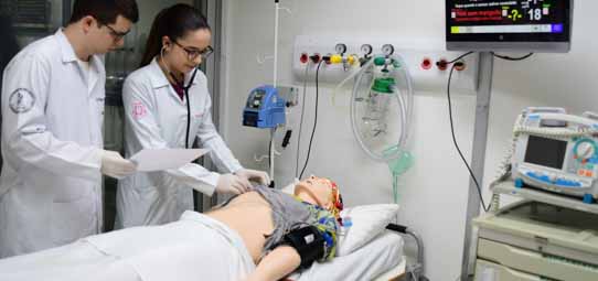 Dois estudantes de Medicina em jaleco branco com brasão institucional e luvas descartáveis realizam procedimentos em manequim de simulação de adulto deitado em maca hospitalar, com monitoramento por eletrodos. Uma estudante usa estetoscópio. O laboratório LHABSIM apresenta monitor com sinais vitais, aparelhos de ventilação e equipamentos hospitalares ao fundo.