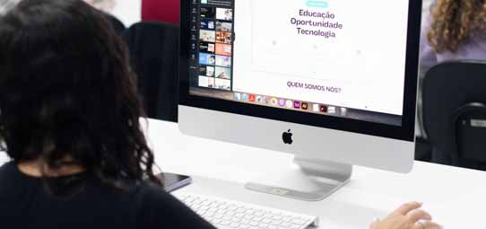 Estudante feminina de costas, com cabelo preto encaracolado, trabalha em um iMac da Apple exibindo na tela uma apresentação com as palavras "Educação, Oportunidade, Tecnologia" e a pergunta "QUEM SOMOS NÓS?". Na barra de tarefas do Mac, ícones de programas de design e edição são visíveis.