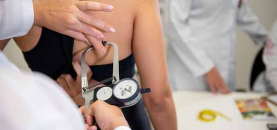 Procedimento de avaliação antropométrica nutricional. Em primeiro plano, mãos enluvadas de um profissional de jaleco branco utilizam um adipômetro (plicômetro) de marca reconhecida para medir a dobra cutânea na região do tronco de uma paciente de top preto. Ao fundo desfocado, outros profissionais de jaleco branco observam o procedimento em ambiente de clínica ou laboratório de Nutrição.