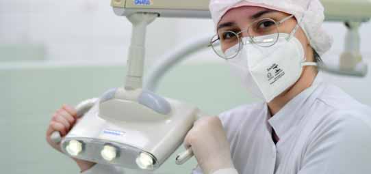Estudante de Odontologia, vestindo jaleco branco, touca rosa, máscara N95 e óculos de proteção, segura e posiciona o foco de luz do equipamento odontológico durante atendimento clínico. O ambiente é uma clínica odontológica com equipamentos modernos. Expressão concentrada e postura profissional.