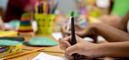 Close nas mãos de um estudante usando caneta hidrográfica preta em folha branca sobre mesa colorida, em ambiente de brinquedoteca ou sala de aula de educação infantil. Ao redor, potes de tinta guache, lápis de cor, réguas e materiais pedagógicos coloridos compõem o cenário de atividade artística.
