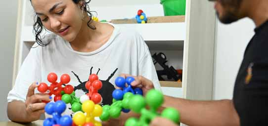 Jovem estudante feminina de camiseta branca com estampa, cabelo cacheado curto, sorri enquanto manuseia um modelo molecular colorido — com esferas vermelhas, azuis, verdes e amarelas interconectadas — em atividade terapêutica ou lúdica. À direita, outra pessoa de camiseta preta também manipula o brinquedo estruturado. Ao fundo, uma estante com materiais pedagógicos e brinquedos. A cena remete a atividade de Terapia Ocupacional ou Psicologia com uso de recursos lúdicos.