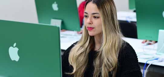 Jovem estudante feminina de cabelo loiro comprido, com batom vermelho e blusa preta, trabalha em computador Apple Mac de gabinete verde em laboratório universitário de Publicidade. Ao fundo, outras estudantes em computadores similares. O ambiente é bem iluminado e organizado.