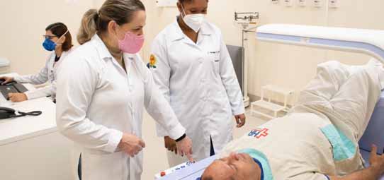 Dois profissionais de saúde com jalecos brancos — uma mulher de máscara rosa e outra de máscara branca — atendem um paciente masculino deitado em maca hospitalar. No fundo, uma terceira profissional de máscara azul trabalha em computador. O ambiente é uma sala de radiologia ou emergência hospitalar clínica.