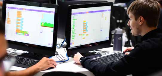 Estudante masculino jovem de camiseta preta trabalha em laboratório de informática com dois monitores Dell à sua frente, ambos exibindo a interface colorida da plataforma Scratch de programação visual em blocos — com blocos laranja, amarelo e roxo e um cenário de jogo com personagem verde. O estudante olha para as telas com expressão concentrada. Uma garrafa de água e teclado preto completam a bancada.