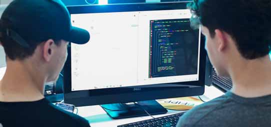 Dois estudantes masculinos de costas trabalham juntos em frente a um monitor Dell de tela dupla exibindo código de programação colorido (sintaxe destacada em verde, amarelo, azul e branco sobre fundo escuro), característico de ambiente de desenvolvimento de software. Um deles usa boné preto. O ambiente tem iluminação azulada, remetendo a laboratório de programação ou hackathon.