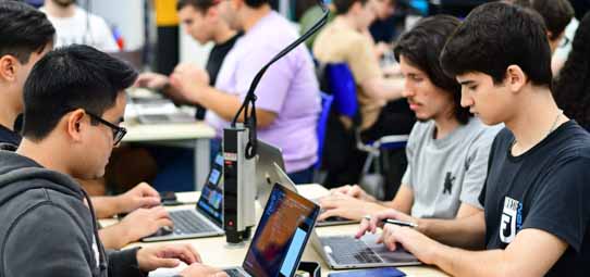 Ambiente de maratona de programação (hackathon) ou laboratório de Sistemas de Informação com múltiplos estudantes jovens trabalhando intensamente em notebooks MacBook e outros laptops dispostos em mesas compridas. Em primeiro plano, dois estudantes masculinos — um asiático de óculos e outro de camiseta cinza — digitam com concentração.