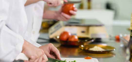 Dois estudantes de Nutrição com dólmã branco trabalham em bancada de aço inox em cozinha profissional. Um deles pica ervas verdes com faca sobre tábua branca, enquanto o outro segura um tomate maduro. Outros tomates, um prato com ingredientes e uma balança de cozinha compõem a cena de aula prática de Nutrição.
